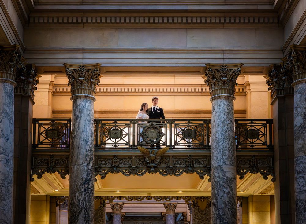 Elegant couple stands on ornate marble balcony with grand columns in a historic capitol building interior.