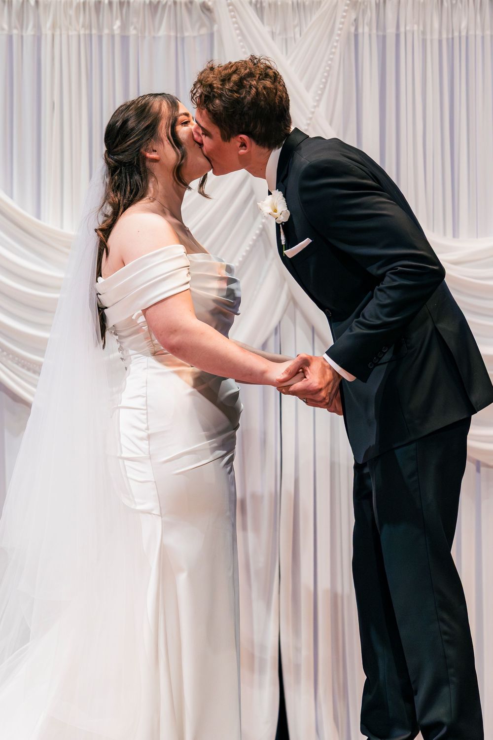 Bride and groom share their first kiss at the altar, dressed in white gown and black tuxedo before draped white backdrop.