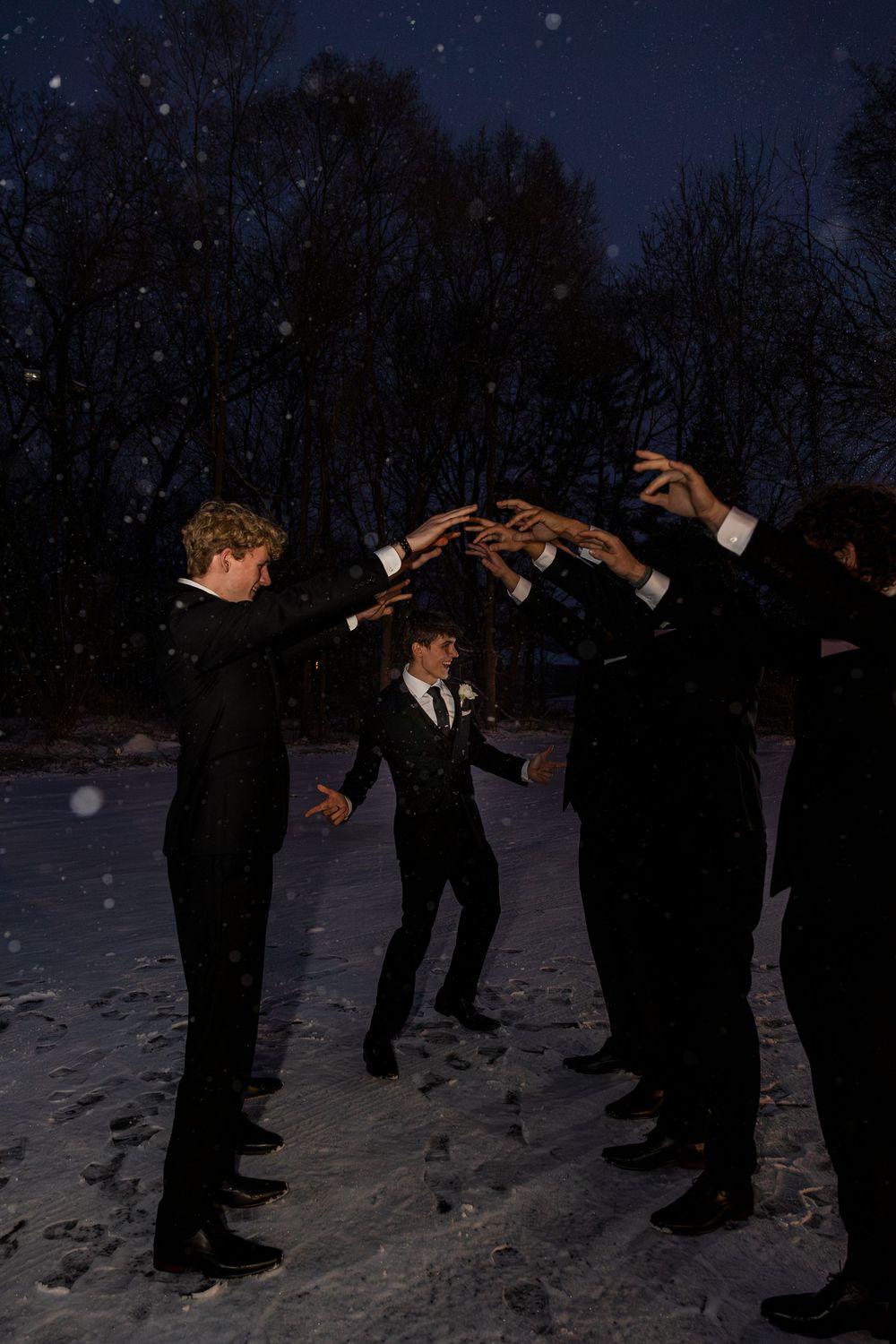 Groomsmen in black suits forming an arch in snowy night setting, with snowflakes falling around them.
