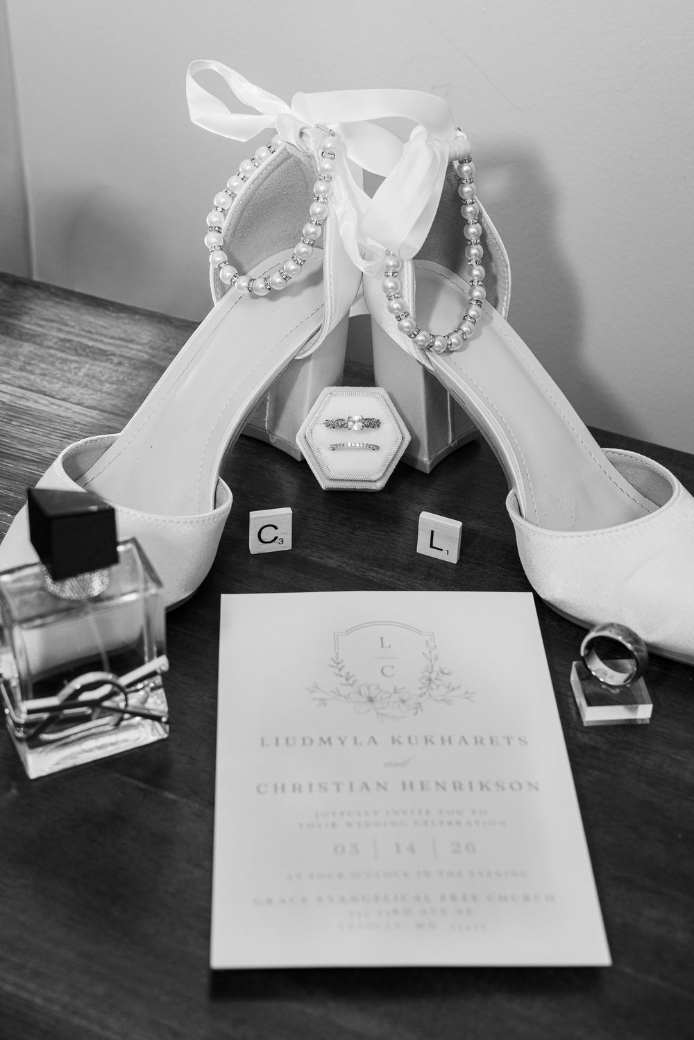 Elegant white bridal shoes and wedding invitation cards arranged on a dark wooden surface in black and white.