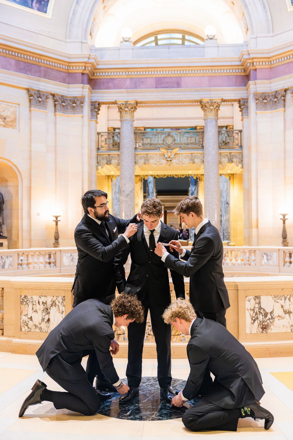 A group in formal black suits gather in a grand ornate room with marble columns and decorative architectural details.