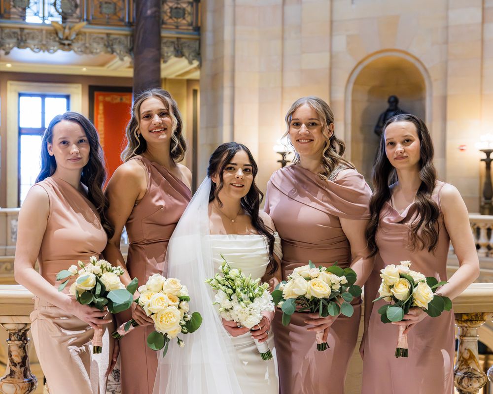 A row of bridesmaids in blush pink dresses hold white floral bouquets while standing in an elegant church setting.