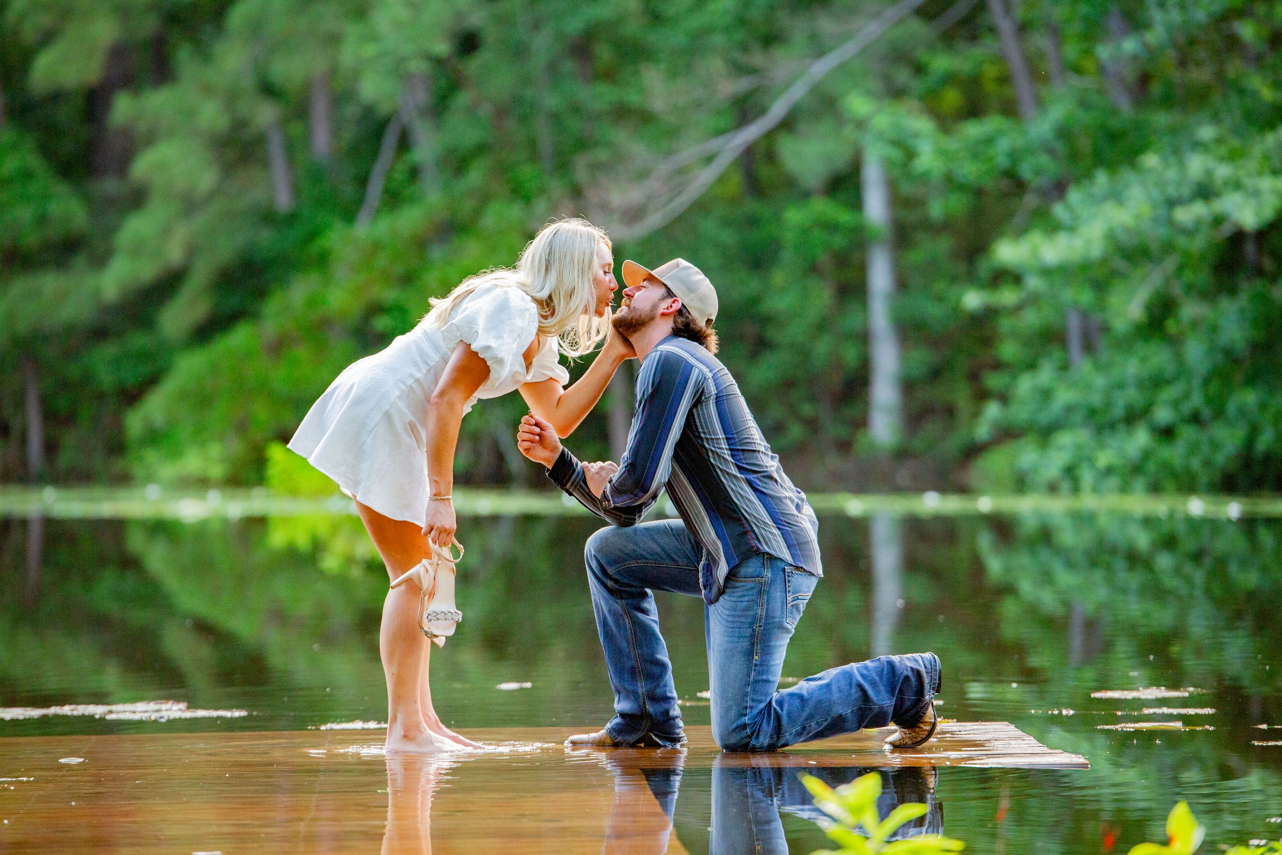 A couple stands together on a dock overlooking a serene lake with green trees in the background.