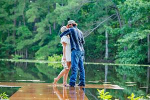 Two people stand close together on a wooden pier with a scenic lake view.
