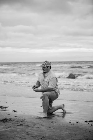 A person in gray kneels on a cloudy beach with waves crashing in the background.