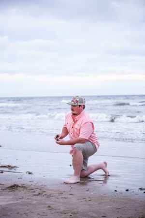 A person in pink kneels on a sandy beach with ocean waves in the distance.