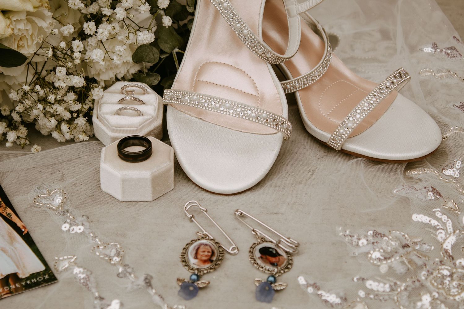 Elegant bridal flat lay featuring crystal-embellished heels, wedding rings, photo charm earrings, and floral accents on a lace surface.
