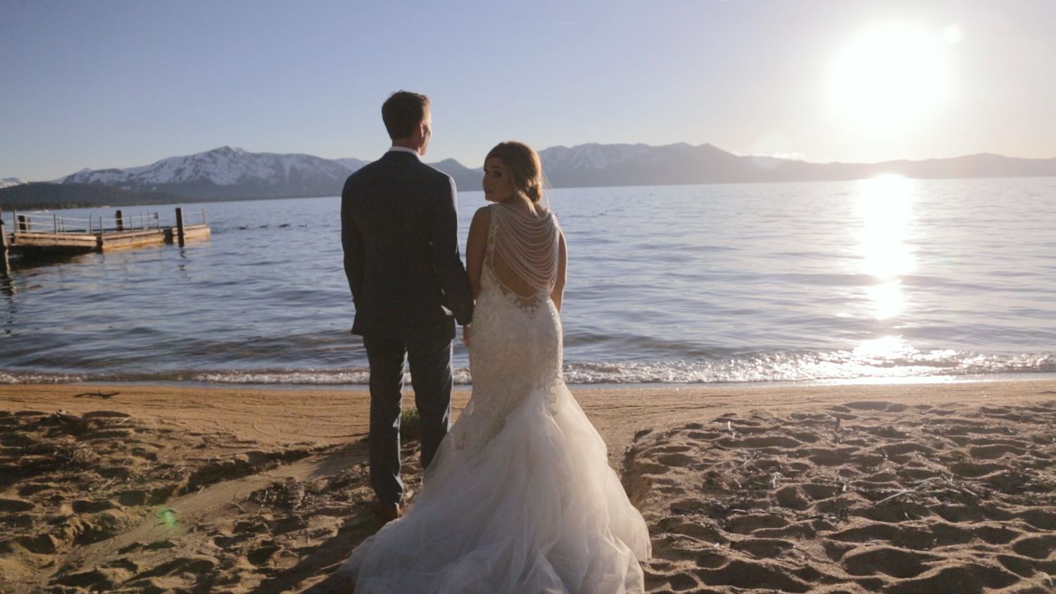 Bride and groom holding hands on sandy beach at sunset with mountain lake views on their wedding day.