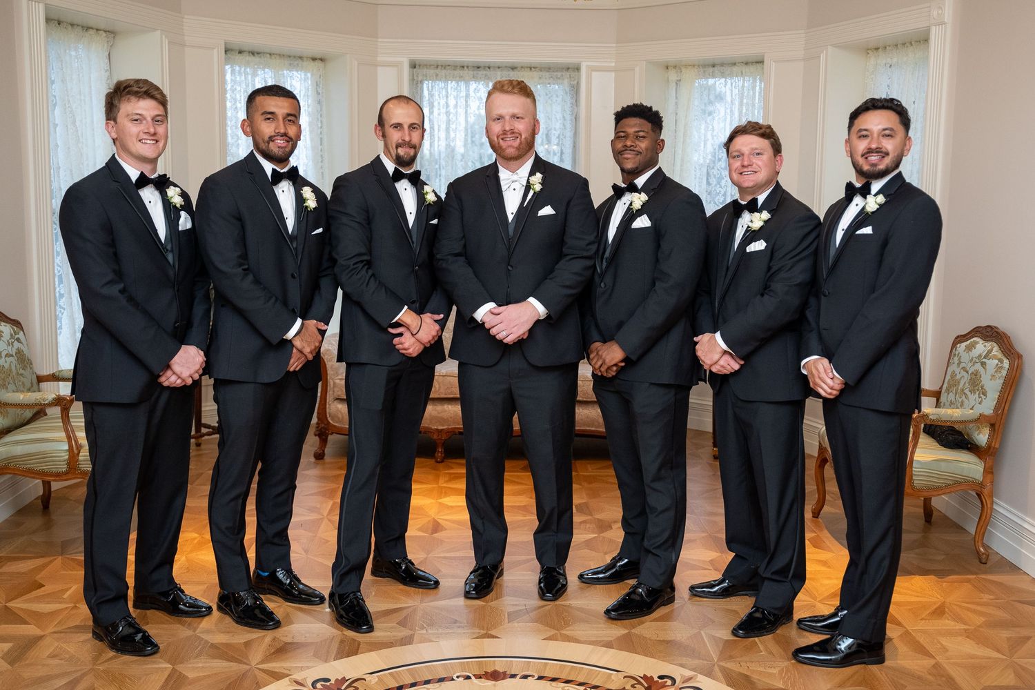A row of groomsmen in black tuxedos stand together smiling in an elegant indoor venue.