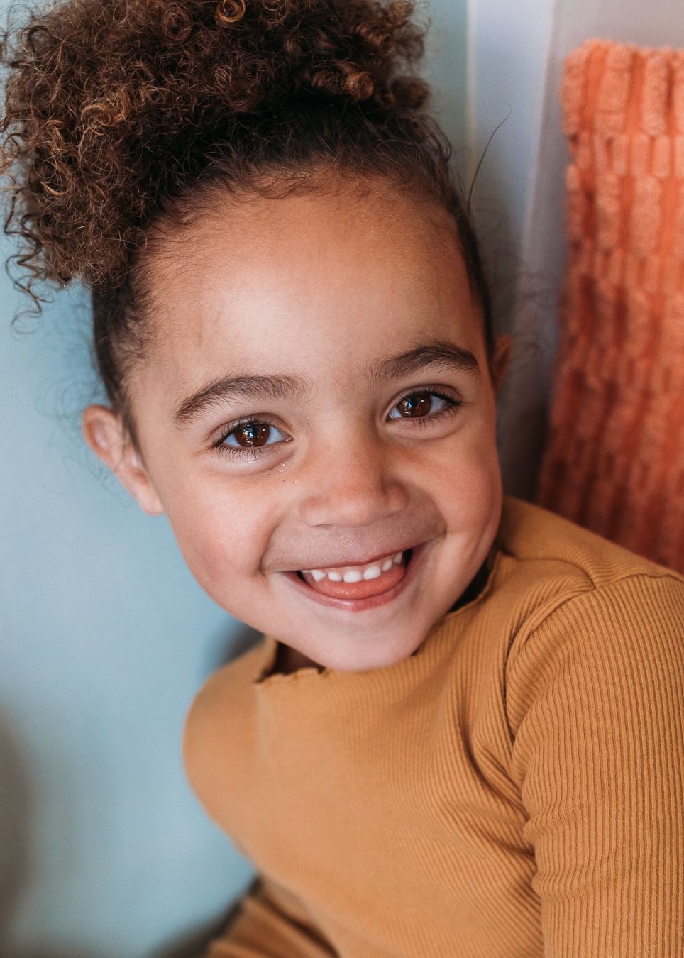 A child wearing a mustard yellow sweater smiles brightly while looking at the camera.
