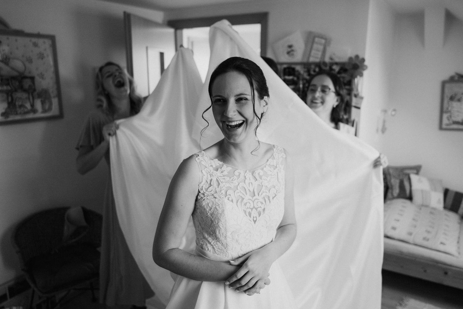 A black and white photo of someone in a delicate lace wedding dress with a flowing veil being held up behind them.