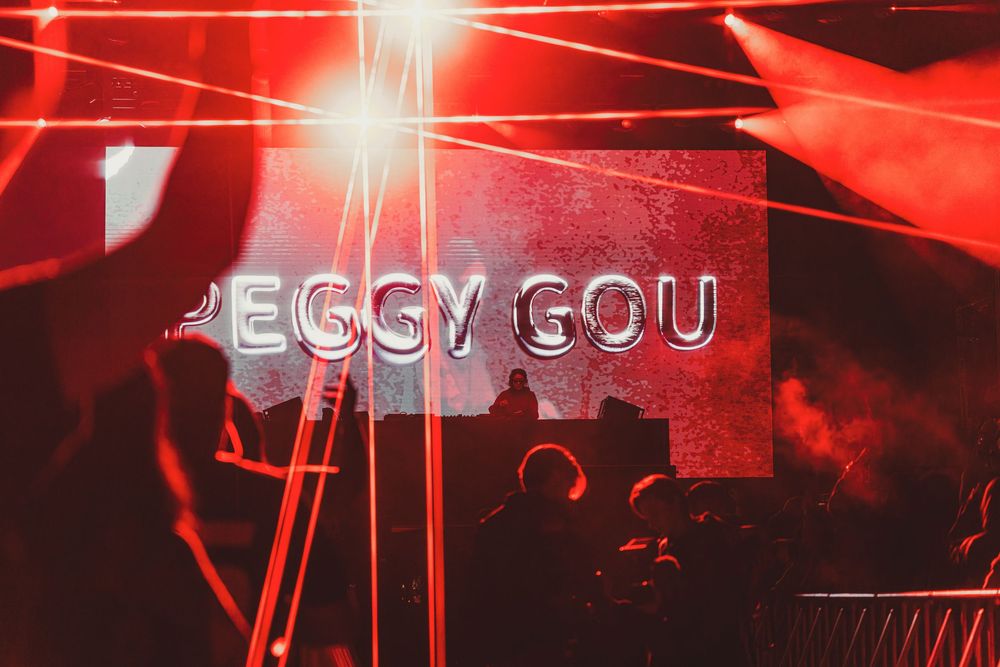 Red concert lighting illuminates a dark stage with bright flares and silhouettes creating a dramatic performance atmosphere.