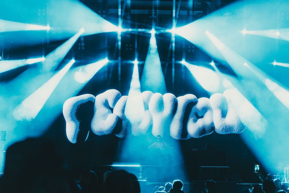 Concert crowd silhouettes with raised hands against dramatic blue stage lighting and beaming spotlights during live performance.