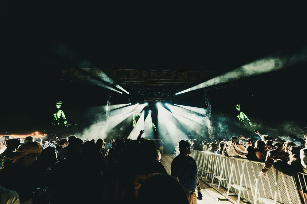Concert crowd silhouettes raise hands in front of bright stage lights during nighttime performance.