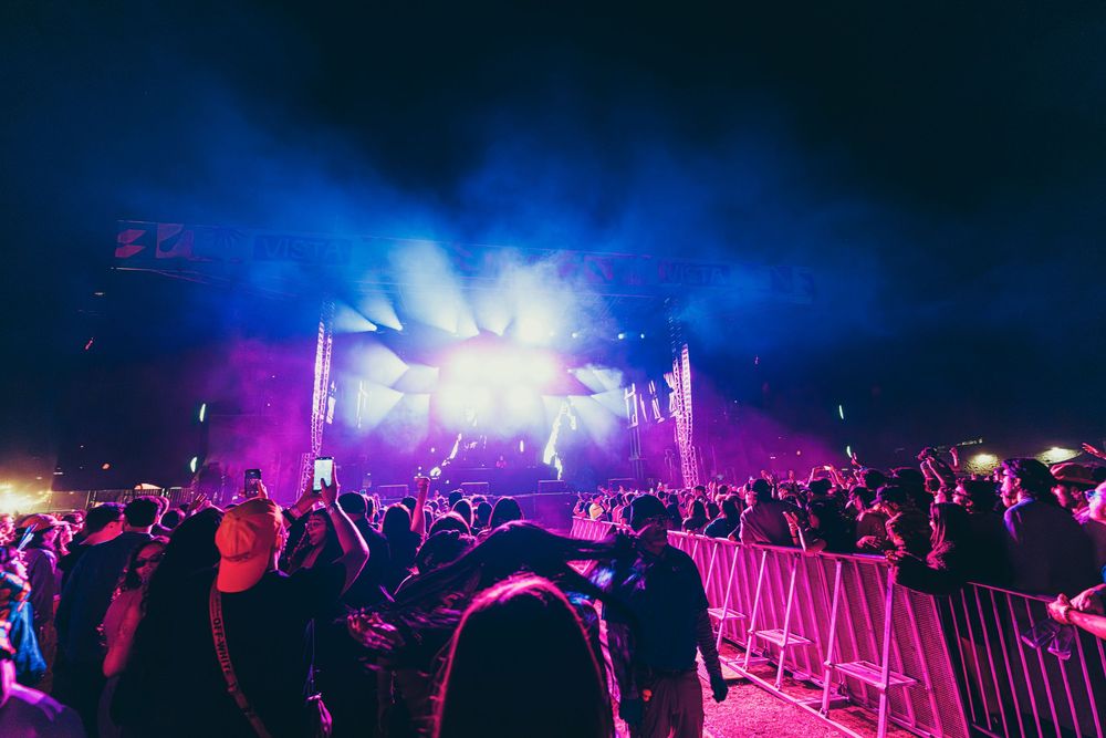 Concert crowd silhouettes facing illuminated stage with bright purple and blue lights during nighttime music festival performance.