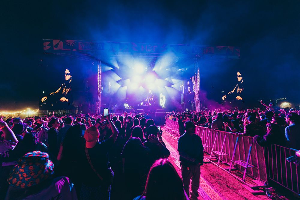 Concert crowd silhouettes against vibrant purple and blue stage lighting at a nighttime music festival performance.