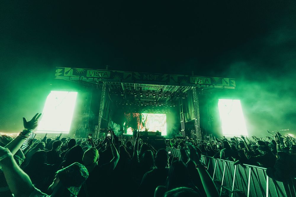 Concert crowd silhouettes raise their hands during a nighttime performance illuminated by bright green stage lighting.