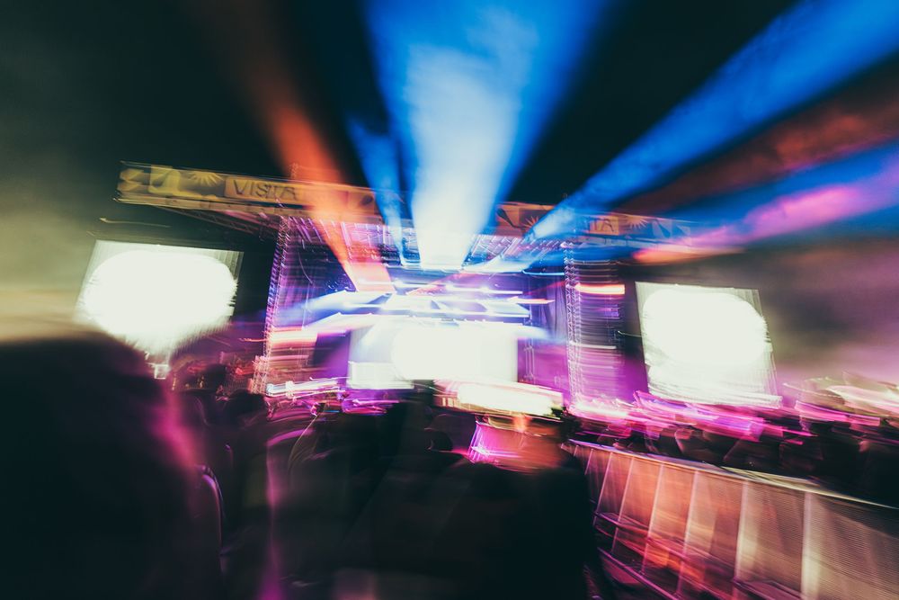 Colorful motion blur of stage lights and LED screens creating dynamic streaks at a concert venue.