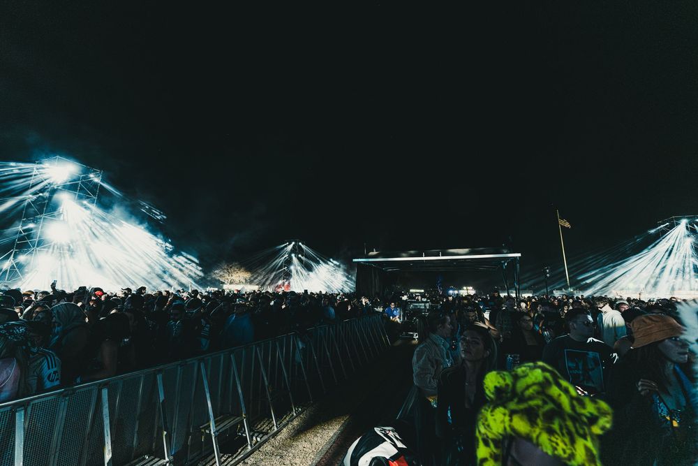Concert venue lit with dramatic white spotlights shining through tent canopy above crowd.