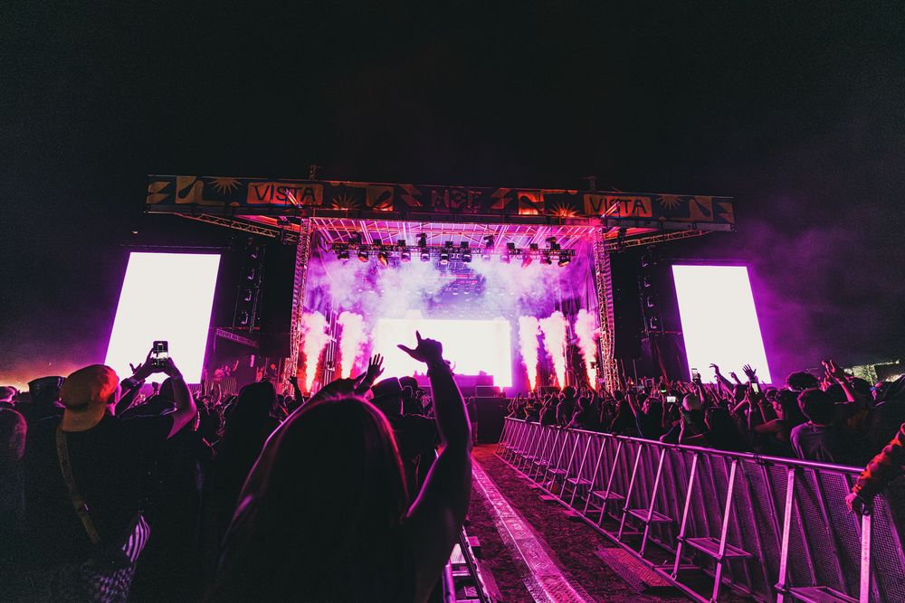 Concert crowd silhouettes against vibrant purple stage lighting at a large indoor music venue during a live performance.
