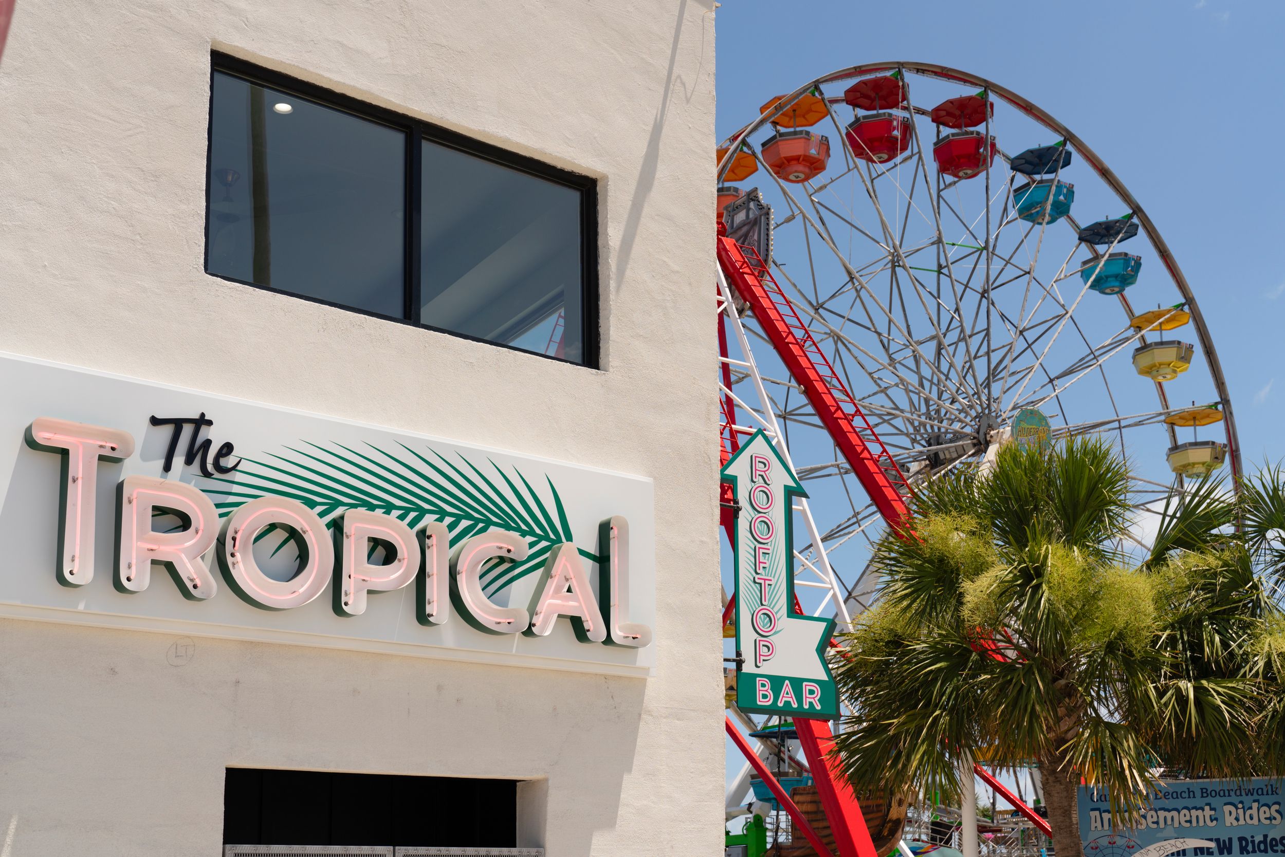 A colorful ferris wheel rises behind The Tropical building sign with palm trees in Las Vegas.