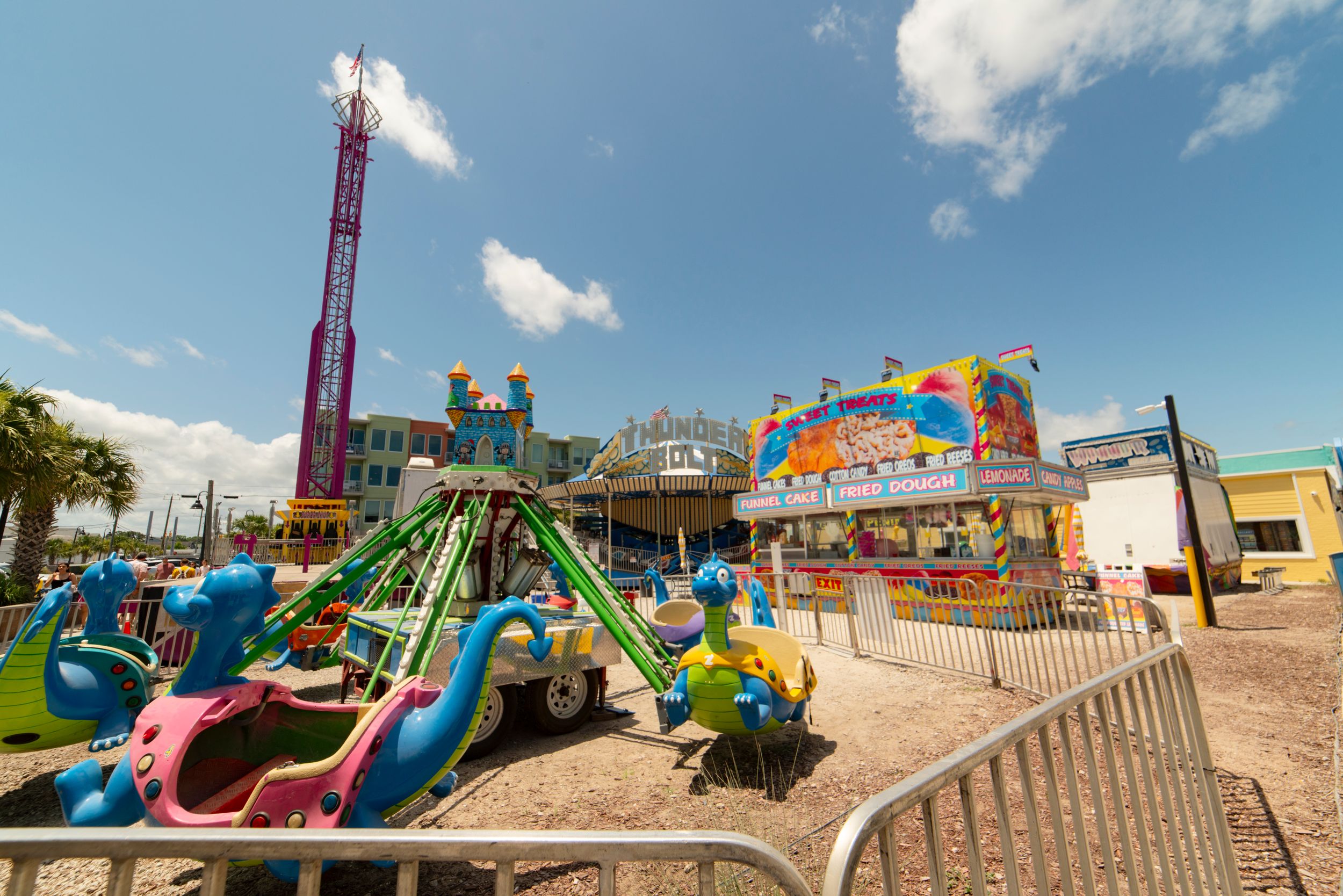 Colorful amusement park rides and attractions under blue sky with tall pink tower and carnival games in background.
