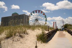 A Ferris wheel and boardwalk path along sand dunes at Myrtle Beach with a hotel building in the background.