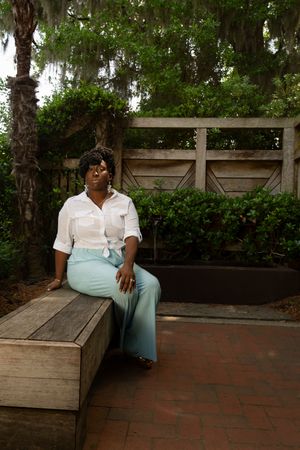 Person in white shirt and mint green pants sits on wooden bench in garden setting with brick paving and greenery.