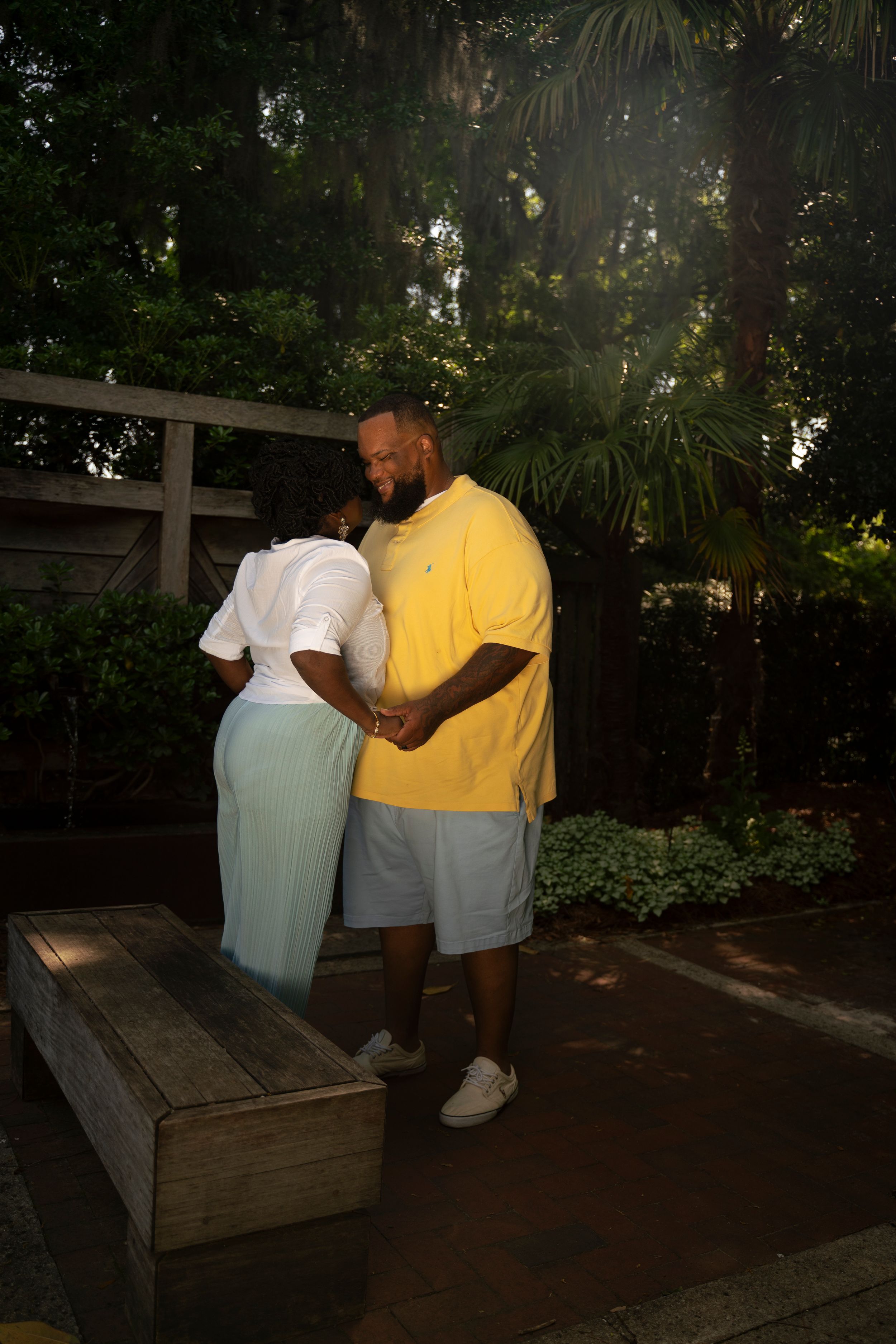 A person wearing a yellow shirt and white shorts tends to a raised garden bed in a shaded outdoor setting.