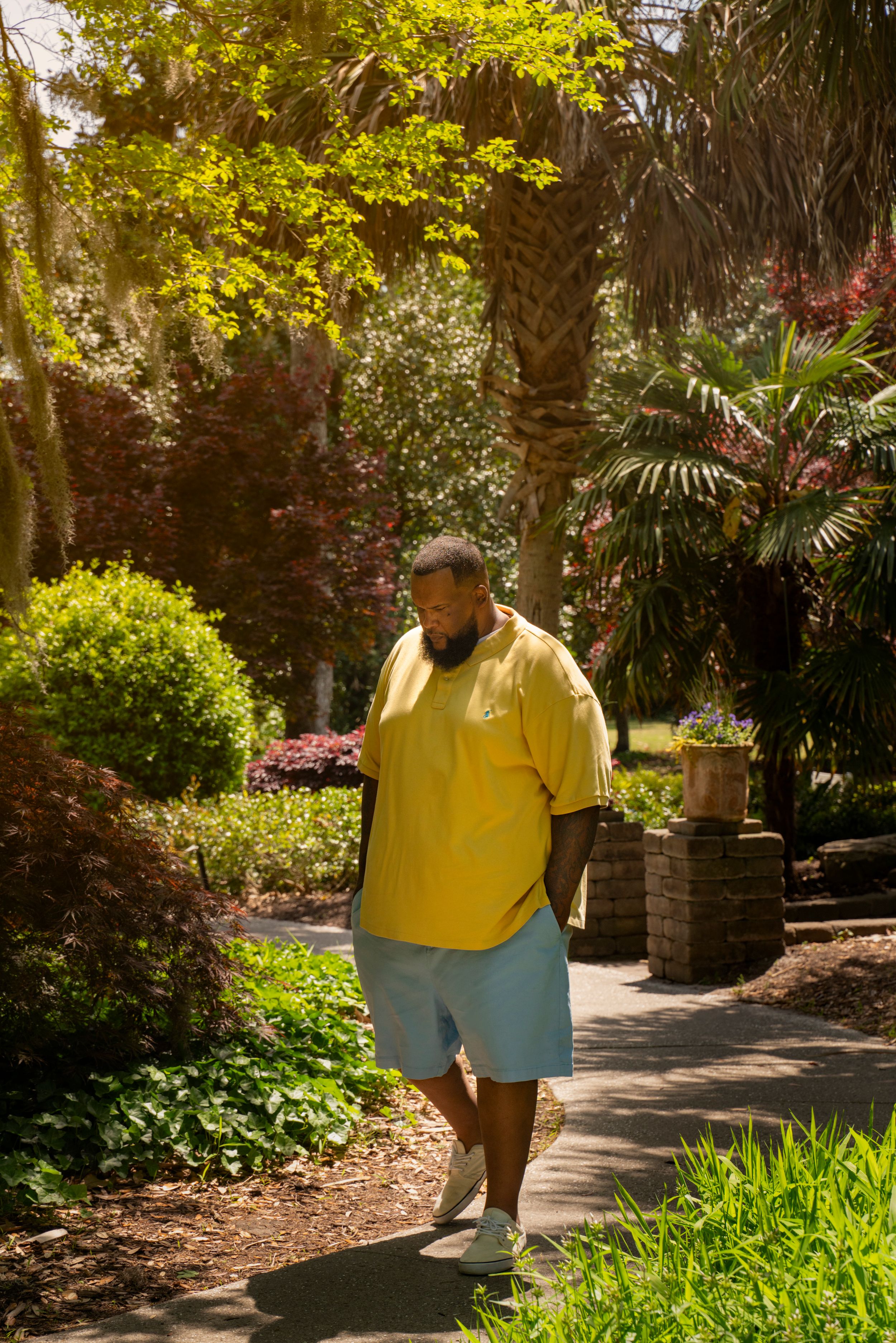 Person in a bright yellow shirt and blue shorts walks along a lush garden path surrounded by tropical plants and palm trees.