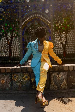 A split-colored blue and yellow suit is worn while posing against a decorative mosaic wall with artistic patterns.
