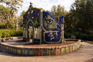 A mosaic wall with colorful tile artwork stretches along a curved garden path in an outdoor park setting.