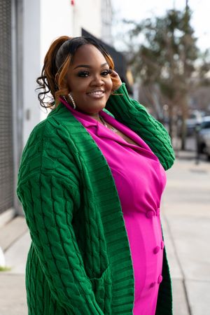 A person poses in a bright green cable knit sweater and hot pink top against an outdoor backdrop.
