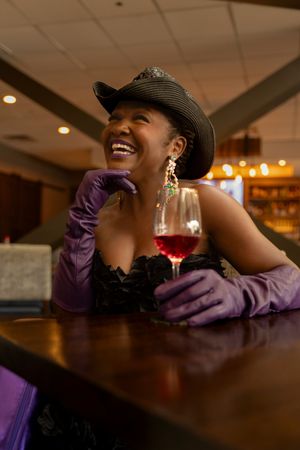 Person wearing purple shirt and black hat laughs while sitting at dark wooden bar with wine glass.