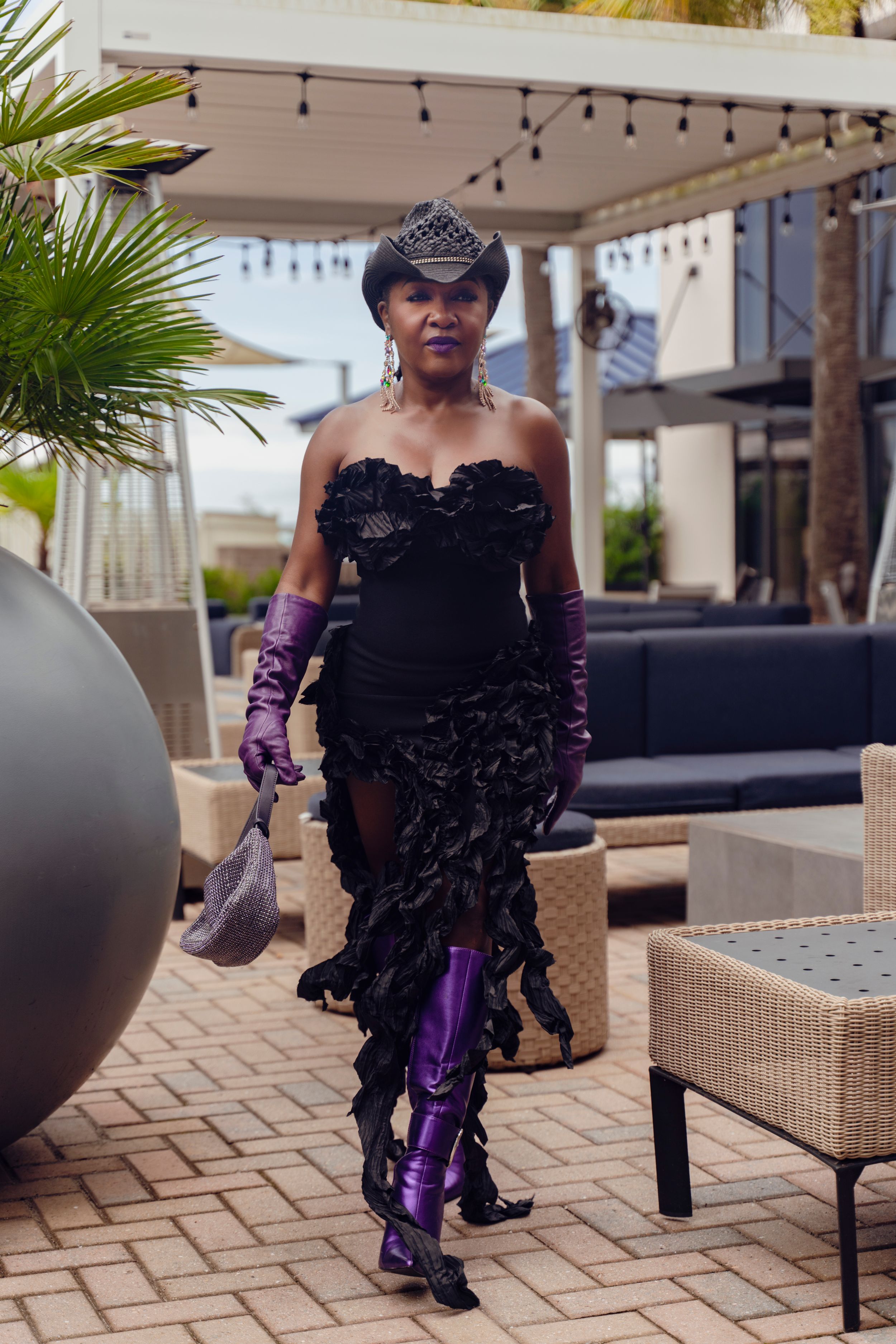 A person poses outdoors in a black lace dress with purple gloves on a patio with wicker furniture and palm trees.