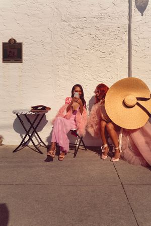 Two people in pink outfits sitting on folding chairs against a white wall with a large decorative hat nearby.