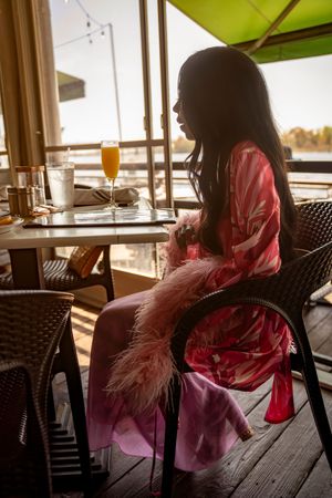 A person in a pink dress sits at a restaurant table near large windows during sunset.