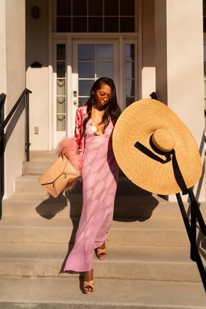 A person in a pink slip dress and jacket stands on steps holding a large straw hat and beige handbag in warm sunlight.