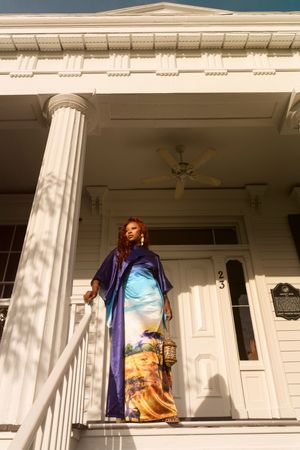 A person in a blue graduation gown poses on a white columned porch with multiple doorways.