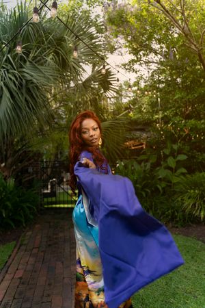 A person wearing a colorful kimono-style dress twirls in a lush garden setting with palm trees and brick pavers.