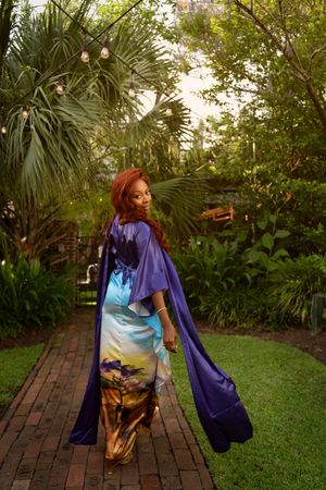 A figure in a purple cardigan and metallic gold pants poses gracefully in a lush garden setting with brick pathways.