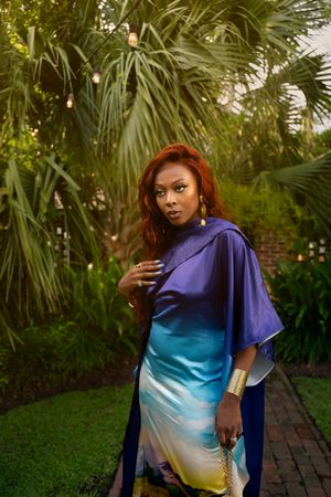 A figure poses gracefully in a flowing gradient dress against a lush tropical garden backdrop with palm trees and brick path.