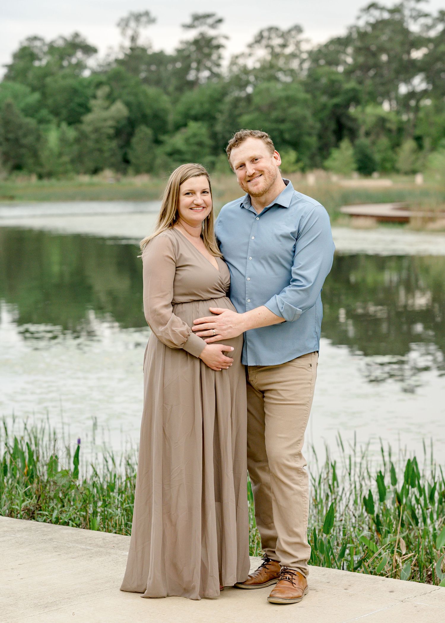 Couple poses together by a serene lake with tall grass and trees in the background during a maternity photoshoot for a houston photographer