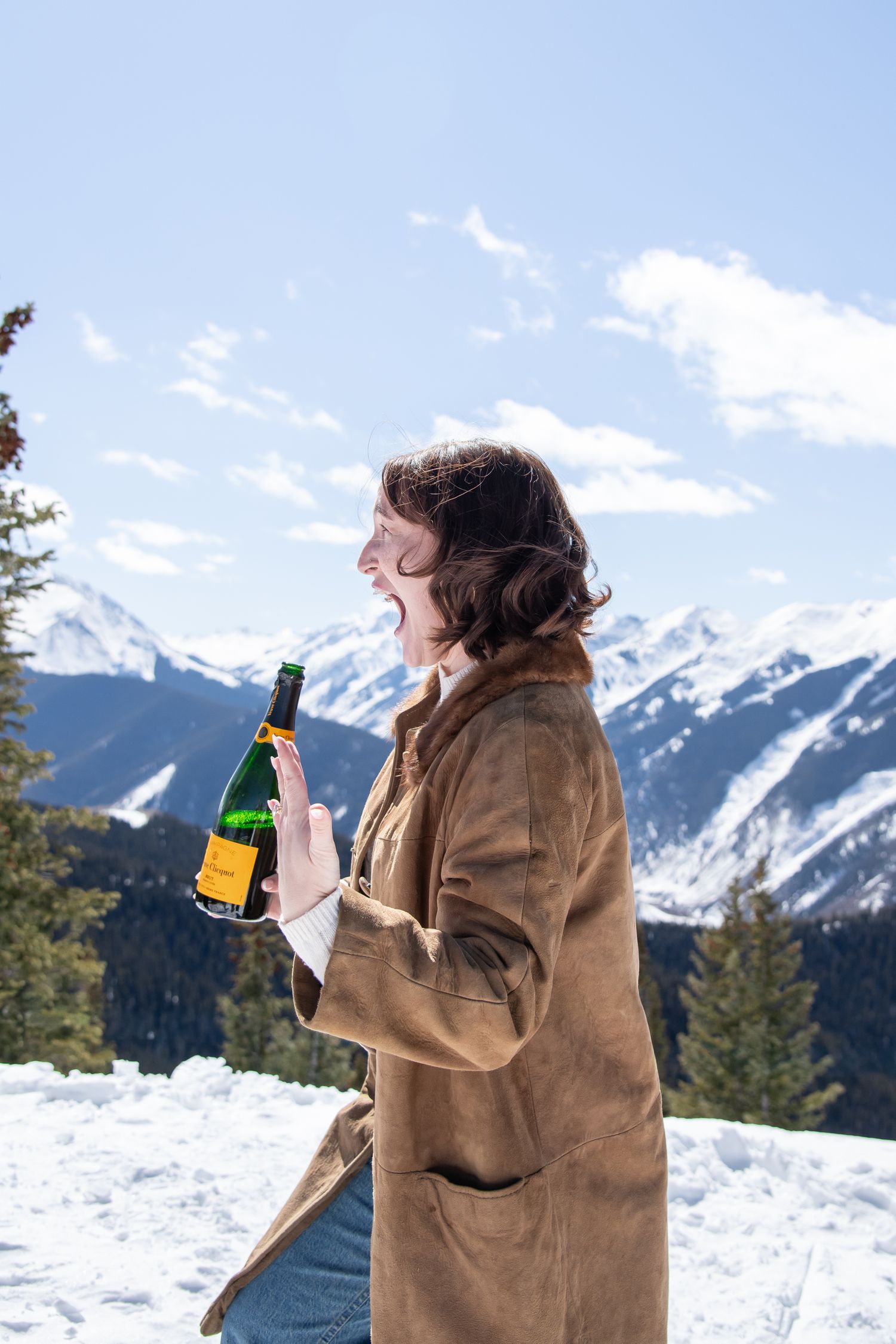 Excited woman show ring and hold bottle of fancy champagne on the top of Aspen mountain during a winter proposal engagement photo session.