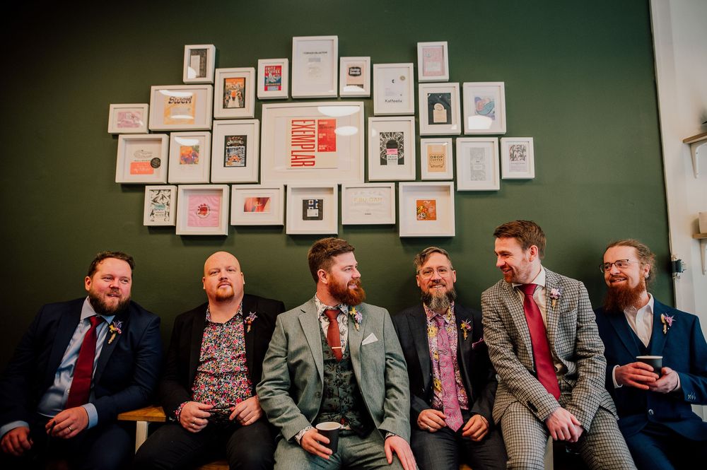 A group of six men in suits sit on a couch laughing together against a green wall decorated with framed artwork.