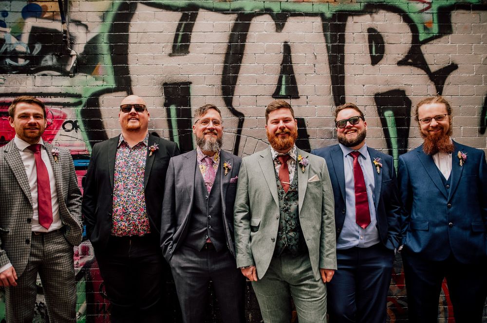 A row of groomsmen in gray suits stand against a rustic brick wall background at a wedding.