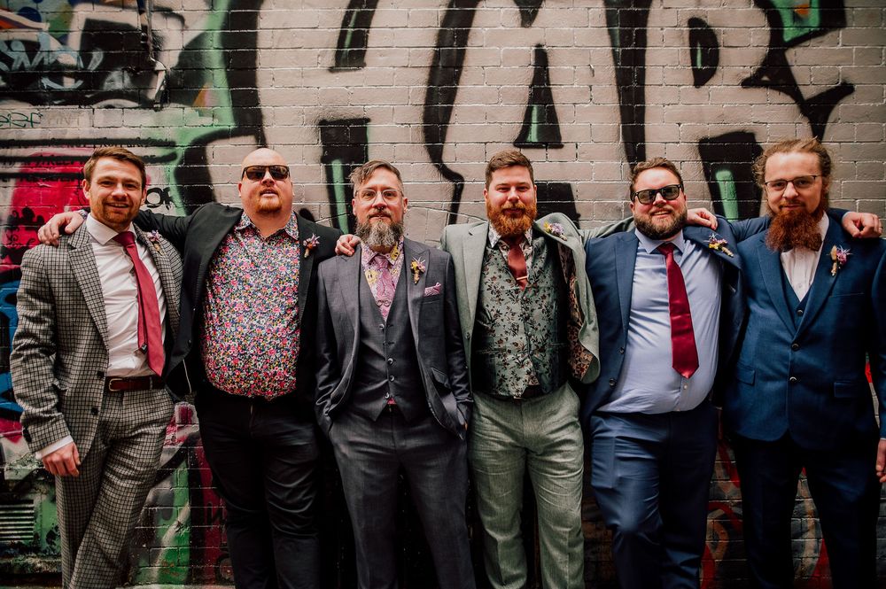 A group of well-dressed men in suits and formal attire pose together for a wedding party photo.