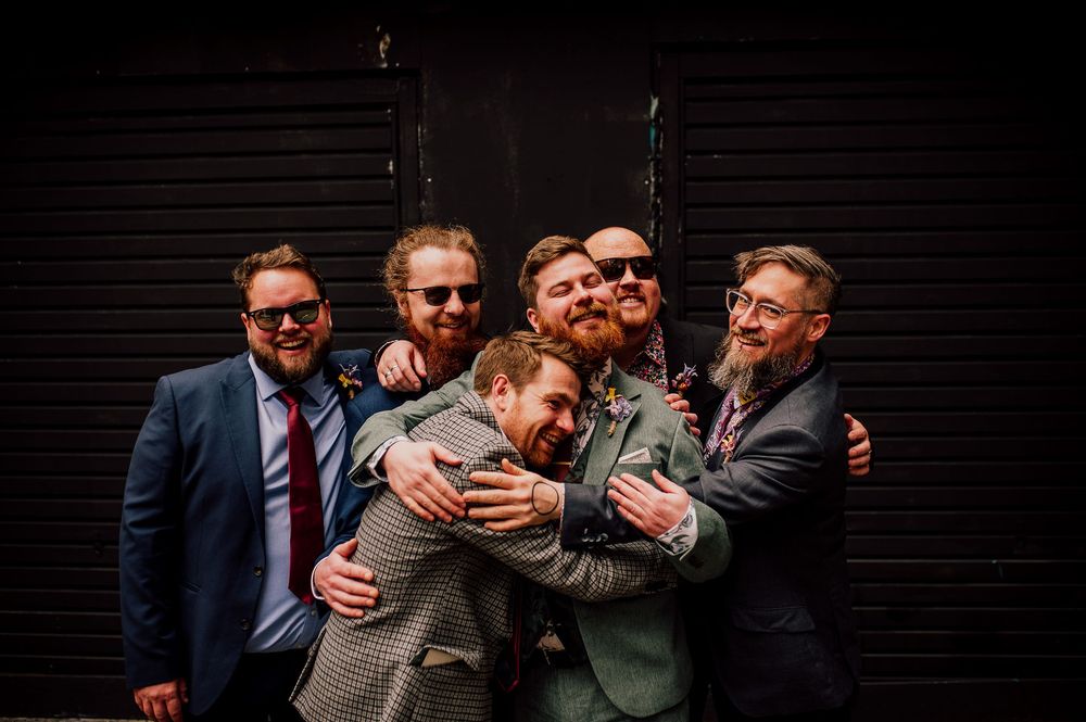A group of groomsmen in suits share a lighthearted moment together against a dark background.