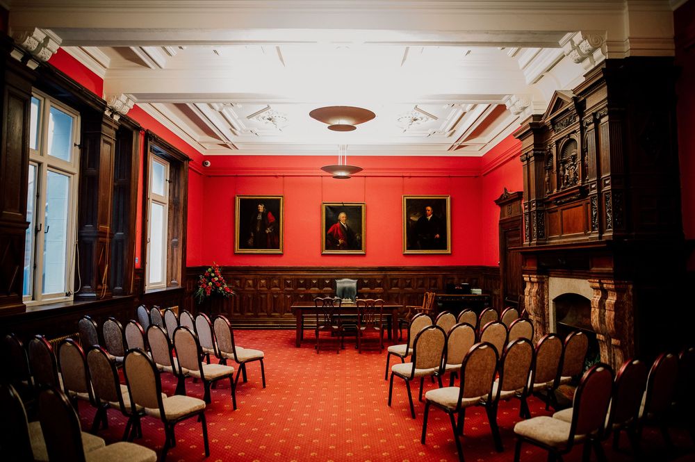 Elegant red meeting room with ornate wood paneling, antique portraits, and rows of upholstered chairs on red carpet.