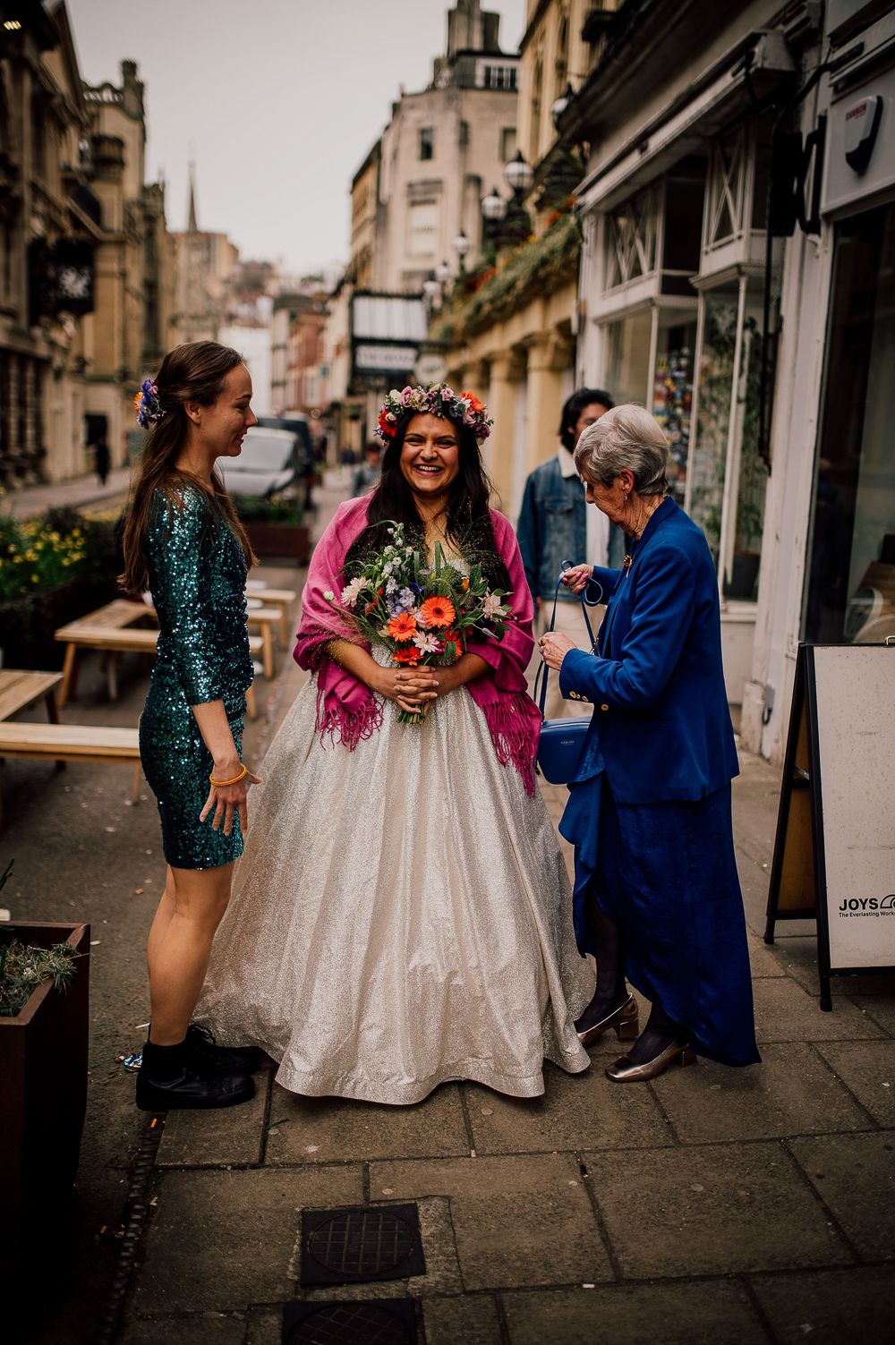 Three people share a joyful moment on a cobblestone street, with one wearing a sparkly dress and another in a white gown.